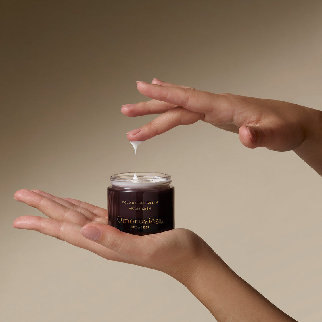 Hands holding Gold Rescue Cream. Picking up a little bit of product with a finger.